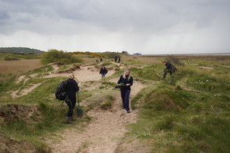 Team members from Cheshire Wildlife Trust walk around bumpy sand dunes on a blustery, overcast day. 