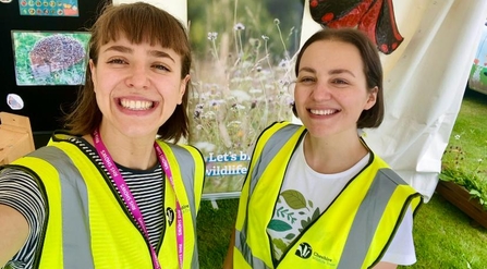 Becky and Rachael setting up for RHS Tatton Flower Show wearing high vis jackets and smiling at the camera.